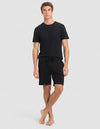 A young man with curly brown hair wears the Cozy Earth Men's Bamboo Stretch-Knit Lounge Tee & Shorts in black. He stands against a light gray background, looking to his left with a neutral expression.|Color:Black