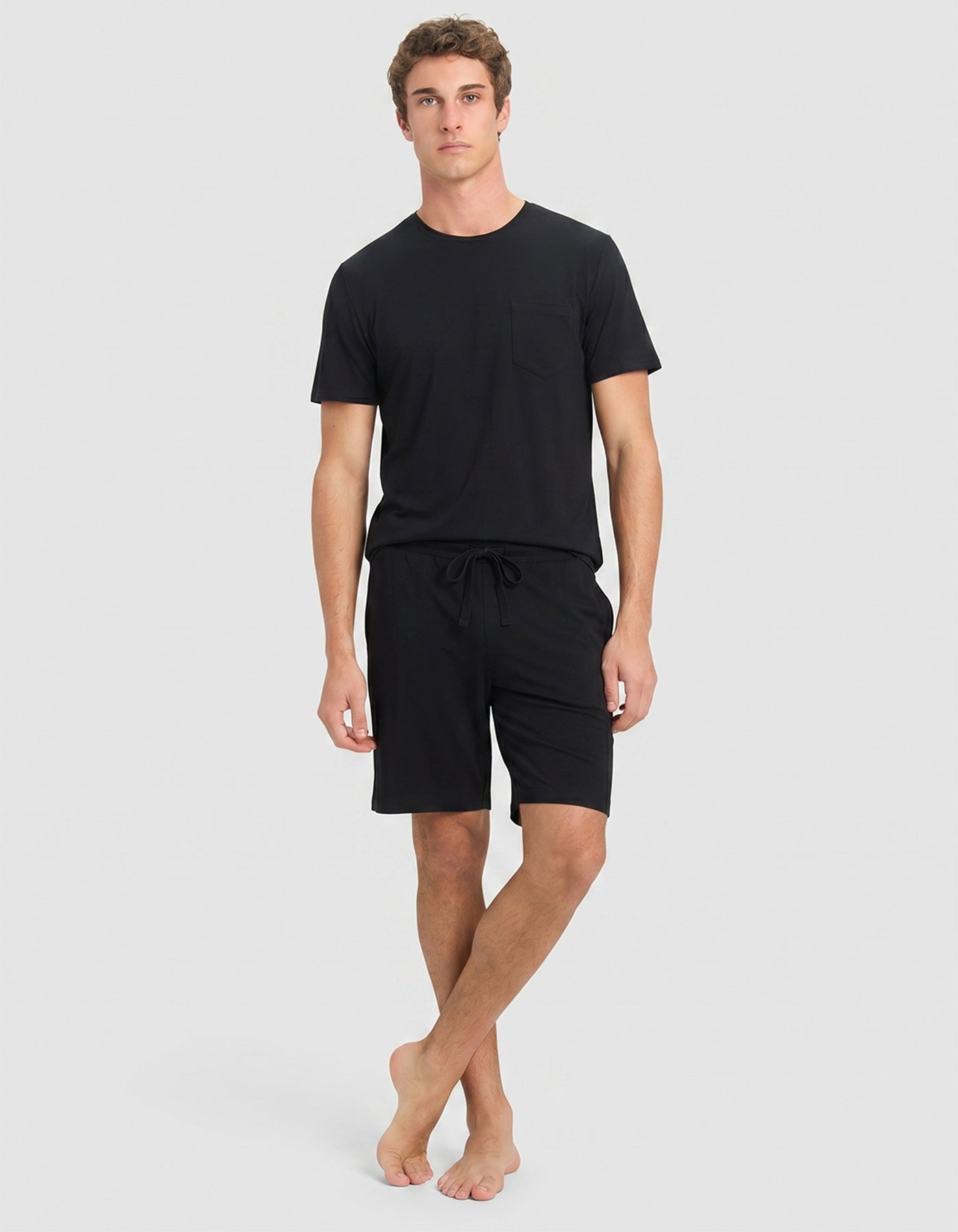 A young man with curly brown hair wears the Cozy Earth Men's Bamboo Stretch-Knit Lounge Tee & Shorts in black. He stands against a light gray background, looking to his left with a neutral expression.