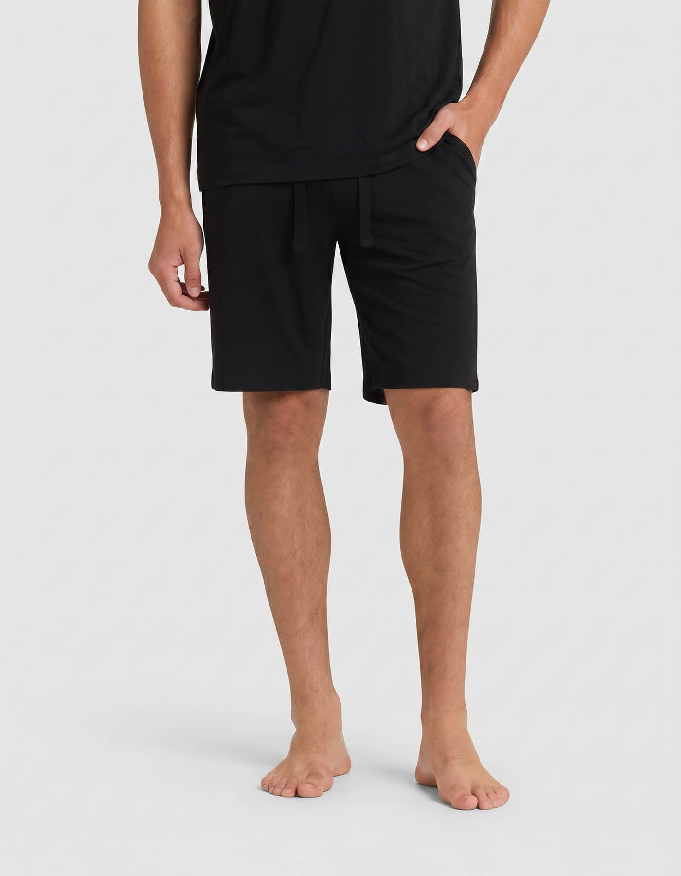 A person stands barefoot against a light gray background, wearing Cozy Earth Men's Bamboo Stretch-Knit Pajama Shorts in black with a black T-shirt. Their hands are relaxed—one in a pocket, the other by their side.