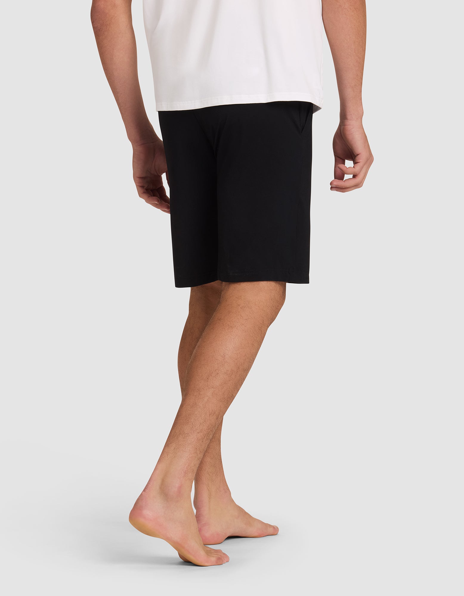 A person stands barefoot on a light gray surface, shown from the back with one leg slightly bent, wearing a white shirt and black Cozy Earth Men's Bamboo Stretch-Knit Pajama Shorts. 