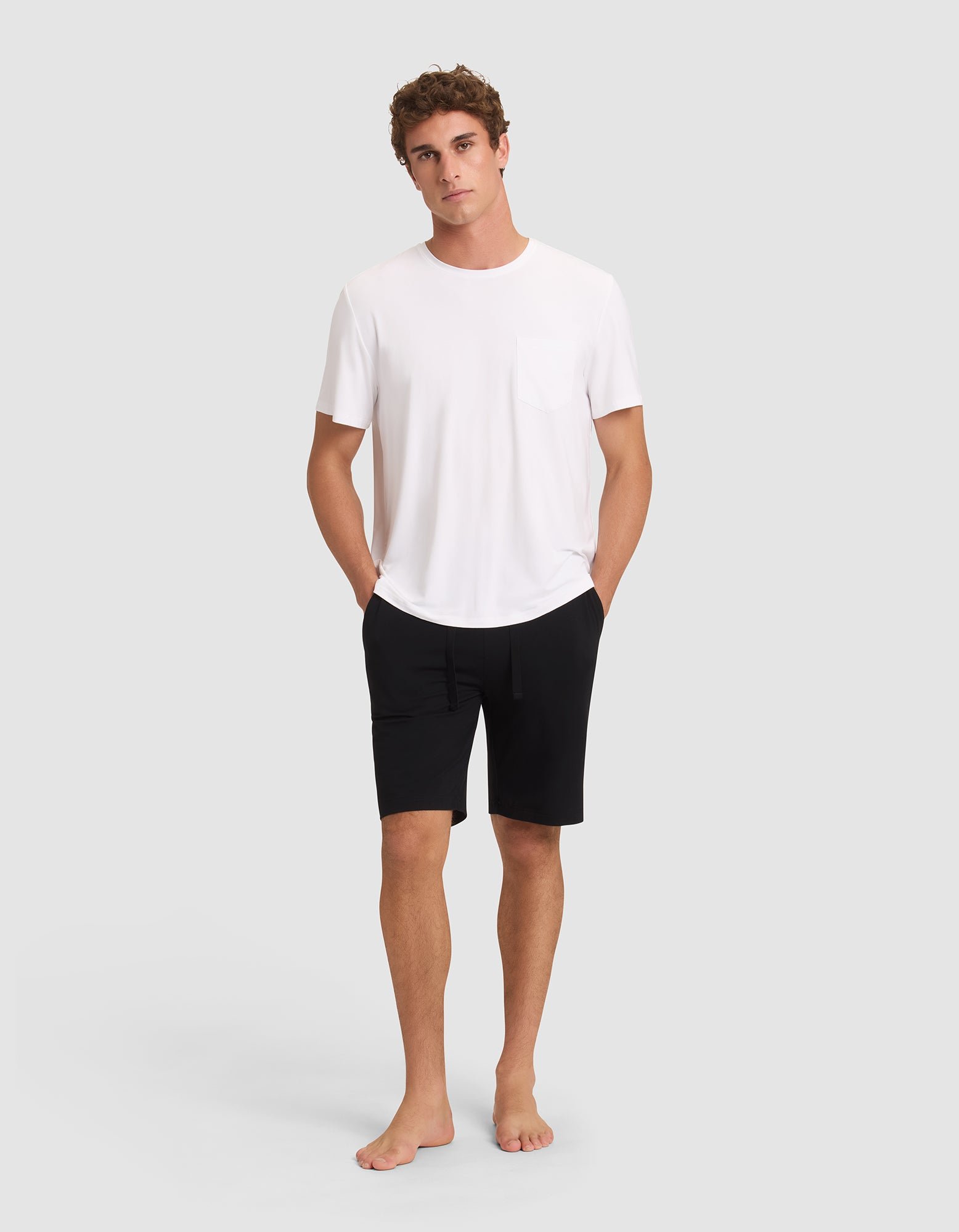 A young man stands barefoot against a plain background, looking relaxed with his hands in his pockets. He wears black Cozy Earth Men's Bamboo Stretch-Knit Pajama Shorts and a white short-sleeve t-shirt with a chest pocket. 