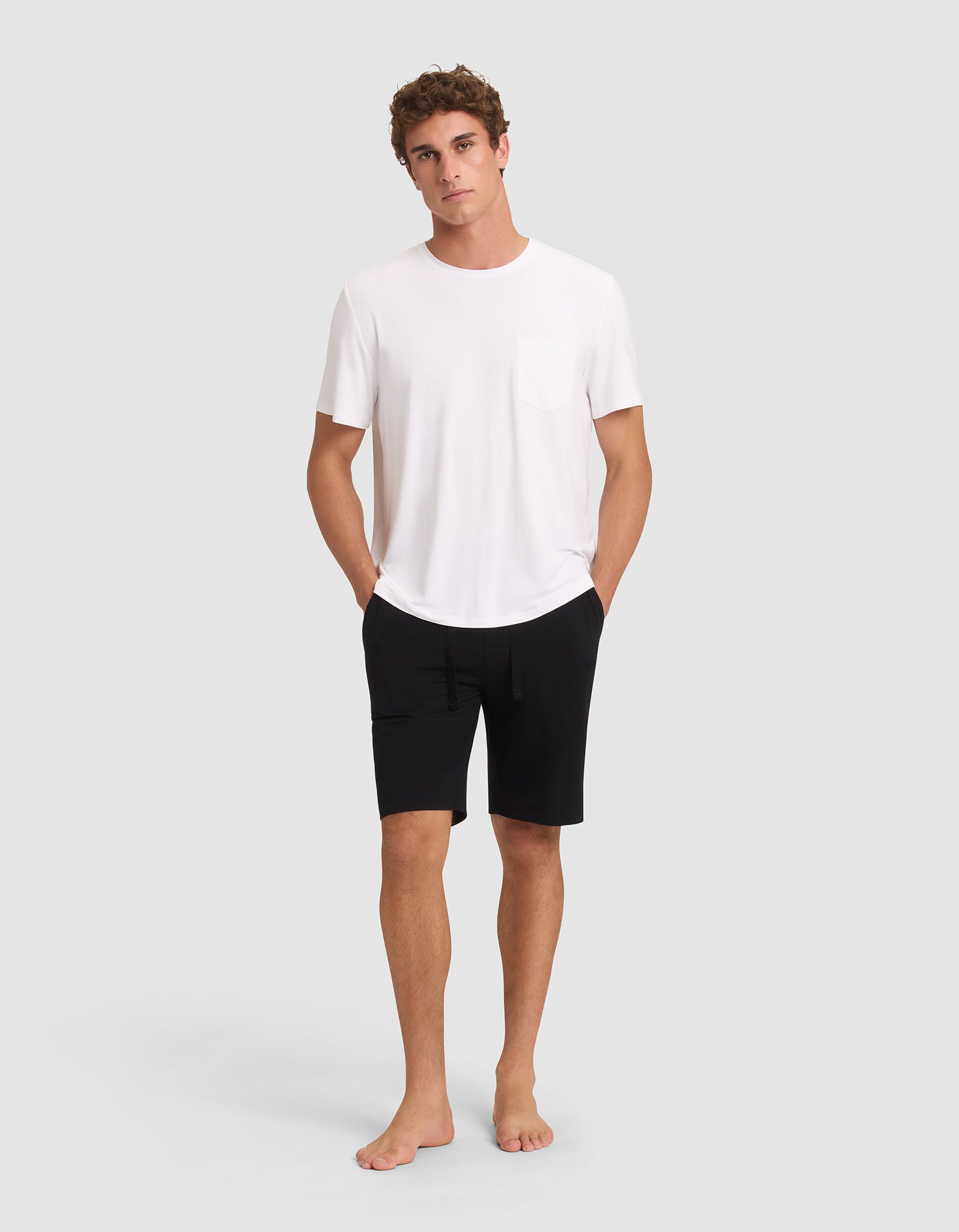 A young man stands barefoot against a plain background, looking relaxed with his hands in his pockets. He wears black Cozy Earth Men's Bamboo Stretch-Knit Pajama Shorts and a white short-sleeve t-shirt with a chest pocket. 