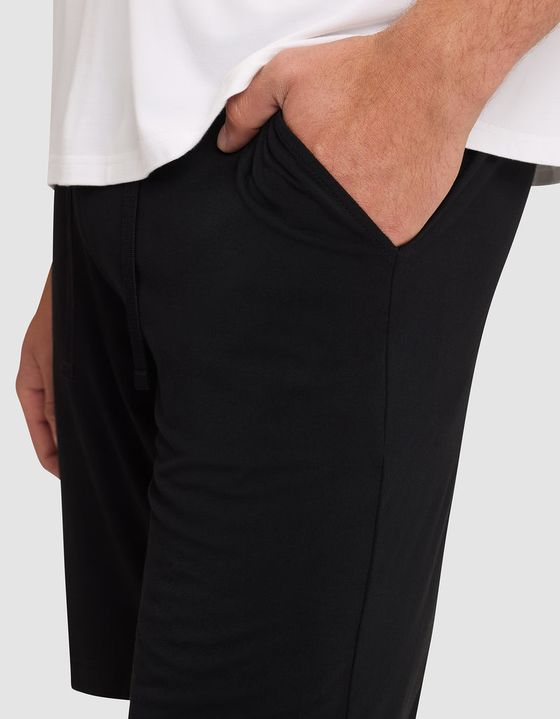 A person is shown from waist to mid-thigh, wearing Cozy Earth Men's Bamboo Stretch-Knit Pajama Shorts in black and a white shirt, with one hand in their pocket.