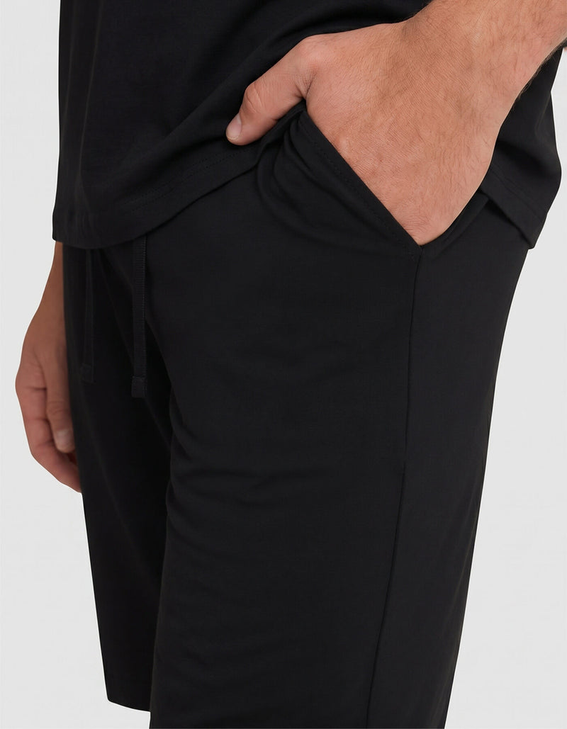 A person is shown from waist to mid-thigh, wearing Cozy Earth Men's Bamboo Stretch-Knit Pajama Shorts in black and a black shirt, with one hand in their pocket.