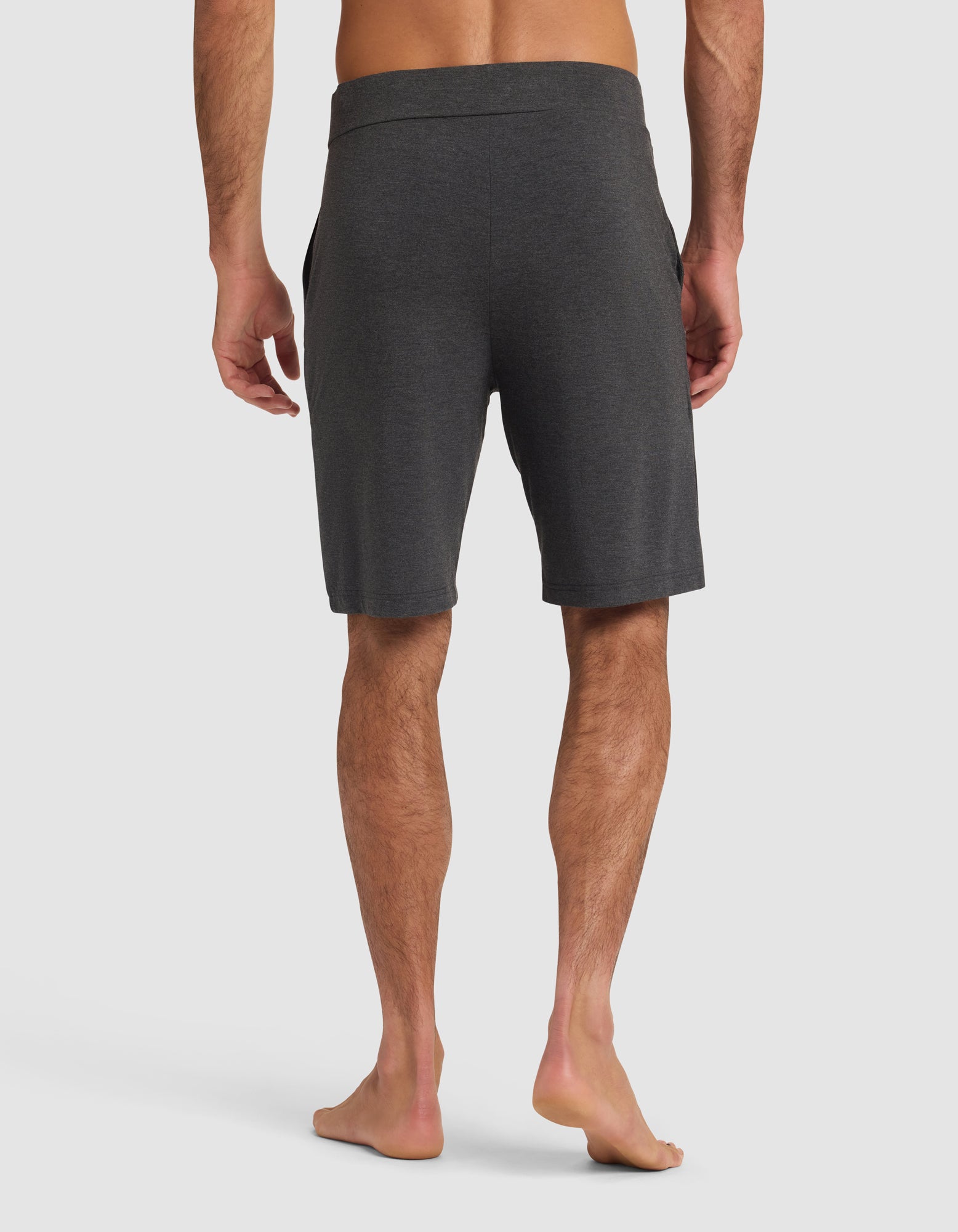 A man stands barefoot facing away, wearing Cozy Earth Men's Bamboo Stretch-Knit Pajama Shorts in dark gray with an elastic waistband; his arms relaxed at his sides against a plain white background.|Color:Charcoal
