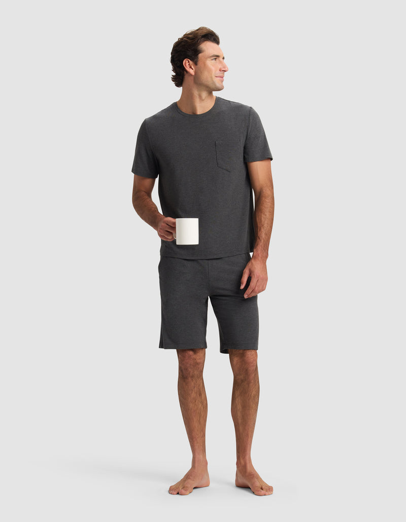 A barefoot man stands holding a white mug, wearing Cozy Earth Men's Bamboo Stretch-Knit Pajama Shorts and a dark gray short-sleeve T-shirt with a chest pocket, set against a plain light background.