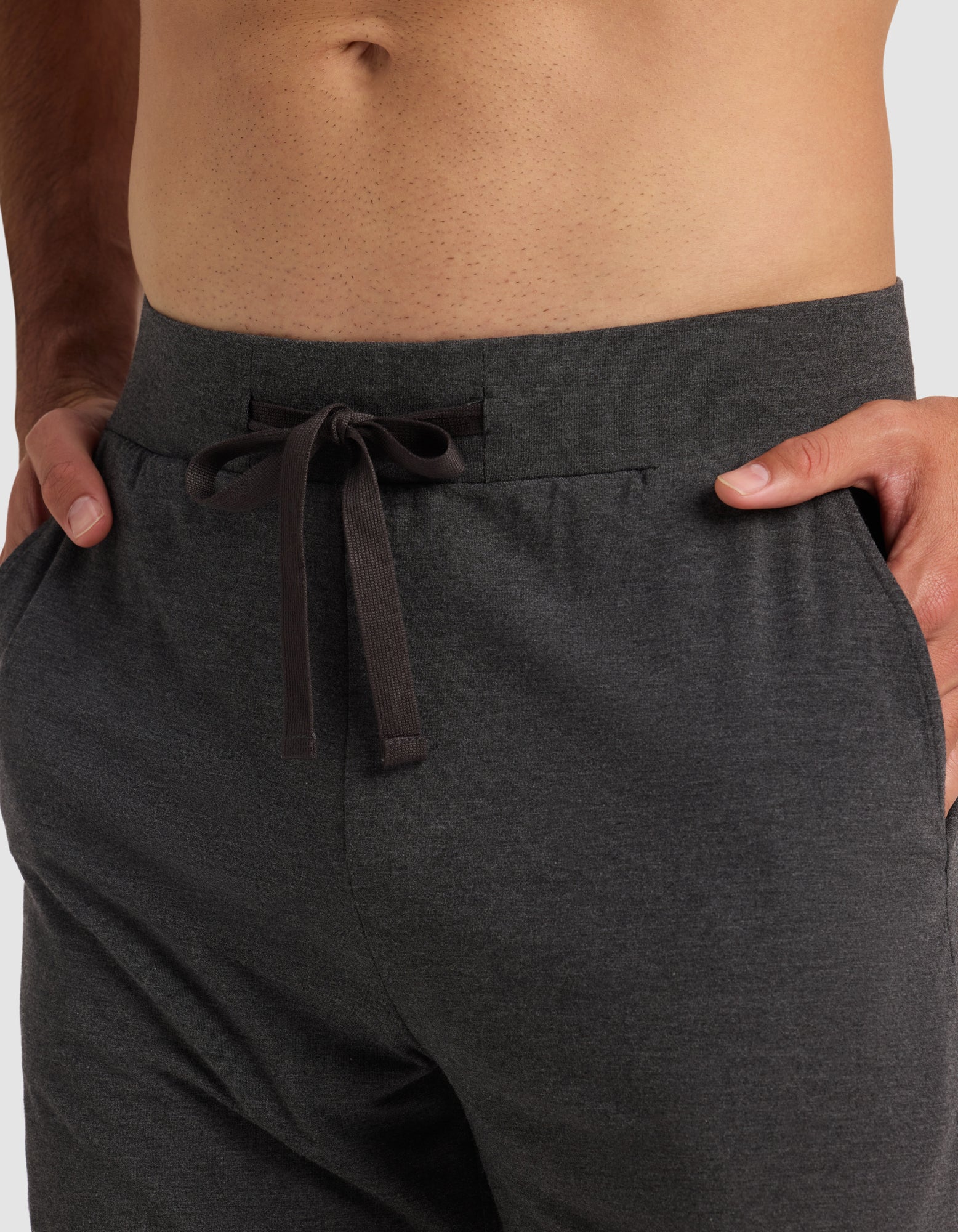 A close-up of a person wearing Cozy Earth's Men's Bamboo Stretch-Knit Pajama Short in dark gray, with hands in the pockets. The shirtless model is shown from waist to upper thighs, highlighting the comfortable stretch-knit fabric.|Color:Charcoal
