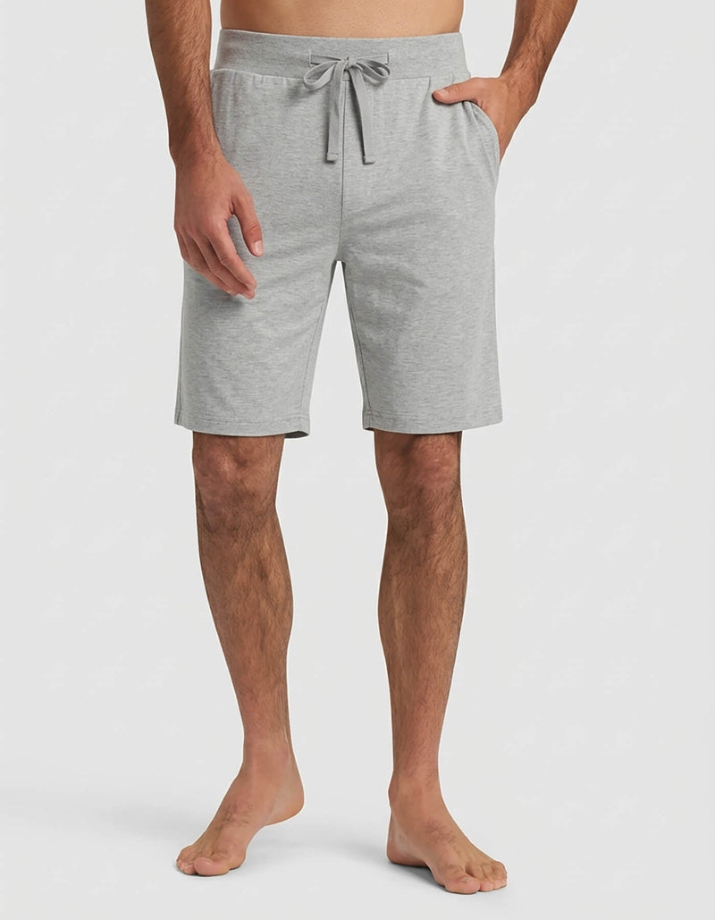 A man wears the Cozy Earth Men's Bamboo Stretch-Knit Short Sleeve Pajama Set in light gray shorts with a drawstring waist and side pockets, standing barefoot against a plain white background.