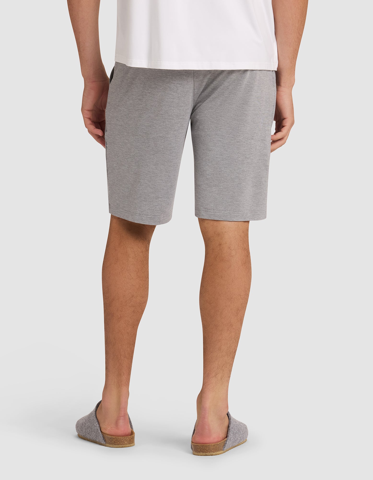 A person wearing Cozy Earth's Men's Bamboo Stretch-Knit Pajama Short in gray, paired with a white shirt and gray slip-on slippers, stands with their back to the camera against a plain background. 