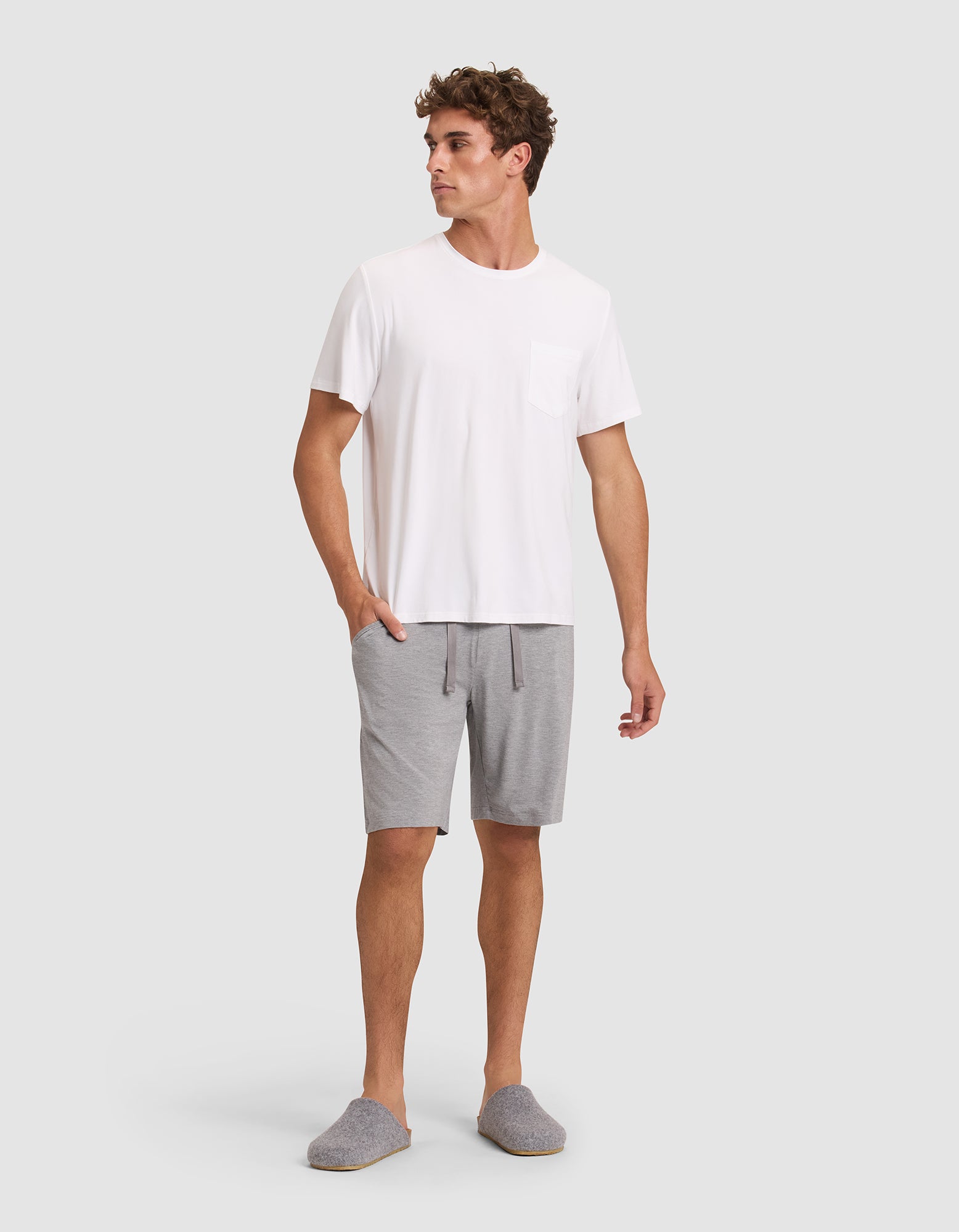 A man stands against a plain background wearing gray slippers, a white tee with a chest pocket, and Cozy Earth Men's Bamboo Stretch-Knit Pajama Shorts in light gray. His left hand is in his pocket and he looks to the side. 