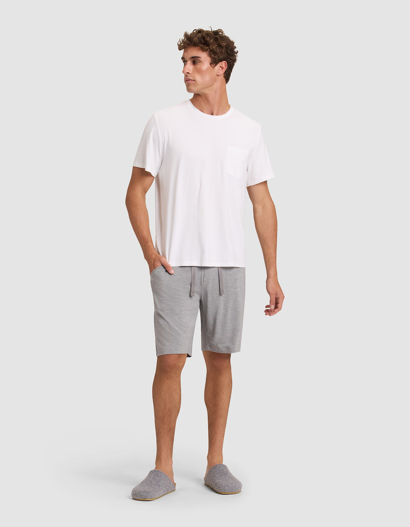 A man stands against a plain background wearing gray slippers, a white tee with a chest pocket, and Cozy Earth Men's Bamboo Stretch-Knit Pajama Shorts in light gray. His left hand is in his pocket and he looks to the side.