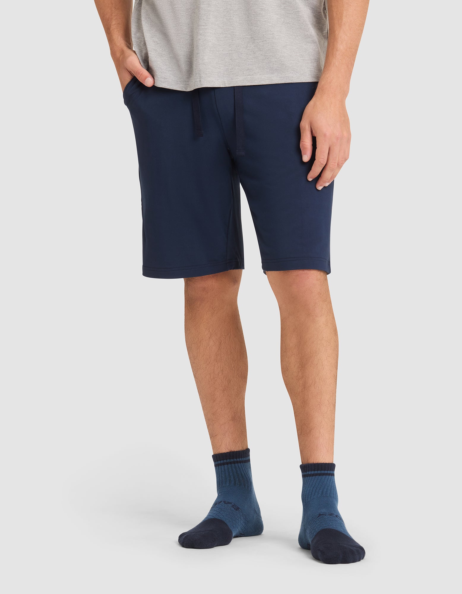 A person stands against a plain light background wearing Cozy Earth's Men's Bamboo Stretch-Knit Pajama Short in navy blue, paired with a gray t-shirt and dark blue socks, with one hand in their pocket. 
