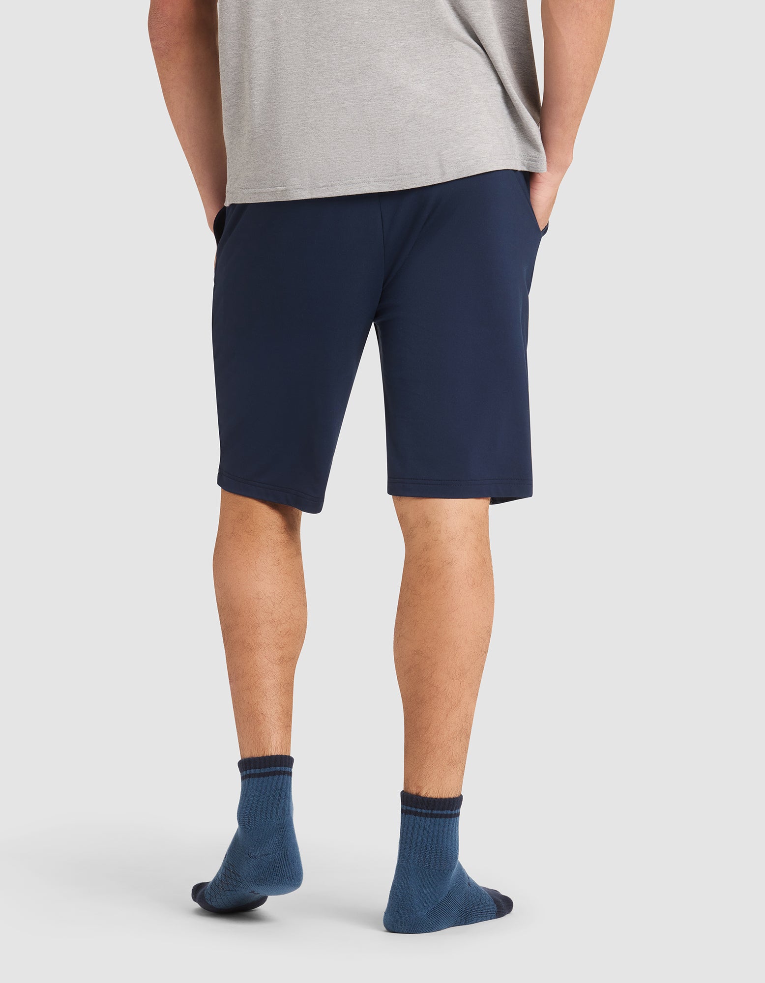 A person stands facing away in a gray shirt and Cozy Earth Men's Bamboo Stretch-Knit Pajama Short in navy blue, paired with blue socks. Their hands are in their pockets against a plain white background. 