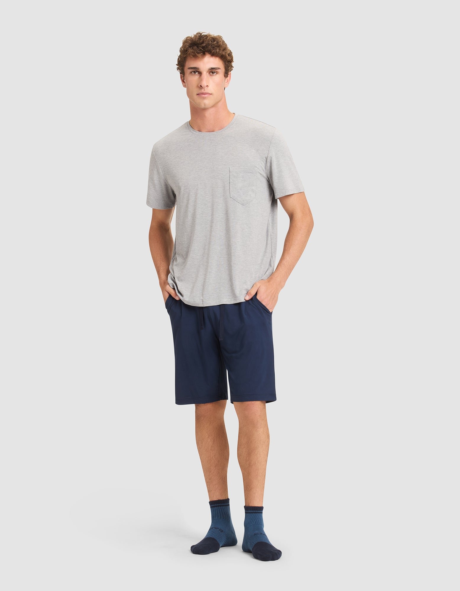 A young man stands against a plain light background, wearing Cozy Earth men's bamboo stretch-knit pajama shorts in navy blue, paired with a light gray T-shirt and dark blue socks. 