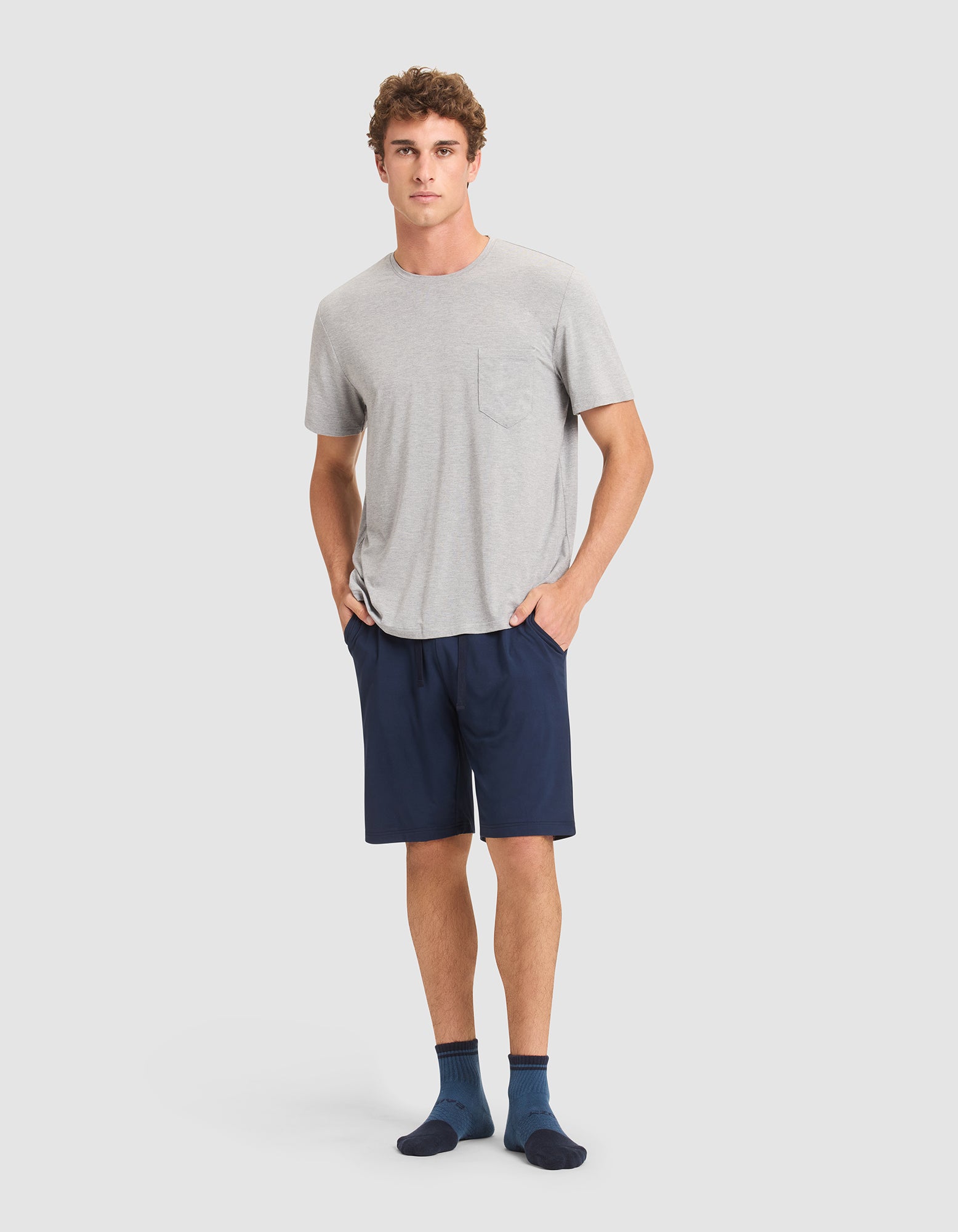 A young man stands against a plain light background, wearing Cozy Earth men's bamboo stretch-knit pajama shorts in navy blue, paired with a light gray T-shirt and dark blue socks. 