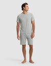 A man stands barefoot against a plain light background, wearing the Cozy Earth Men's Bamboo Stretch-Knit Short Sleeve Pajama Set in Heather Grey with a chest pocket and matching shorts, looking slightly to his left in a relaxed pose.|Color:Heather Grey
