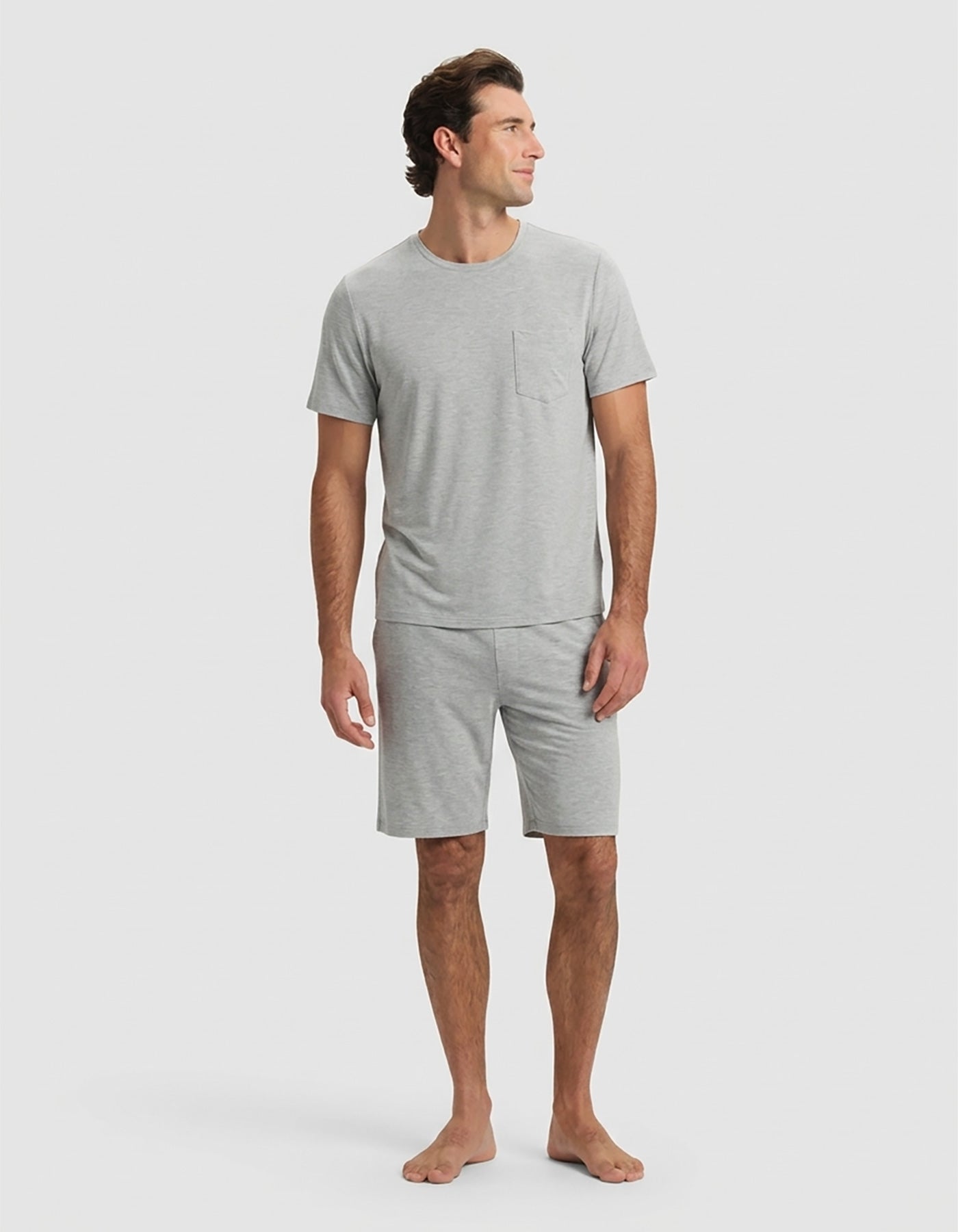 A man stands barefoot against a plain light background, wearing the Cozy Earth Men's Bamboo Stretch-Knit Short Sleeve Pajama Set in Heather Grey with a chest pocket and matching shorts, looking slightly to his left in a relaxed pose.