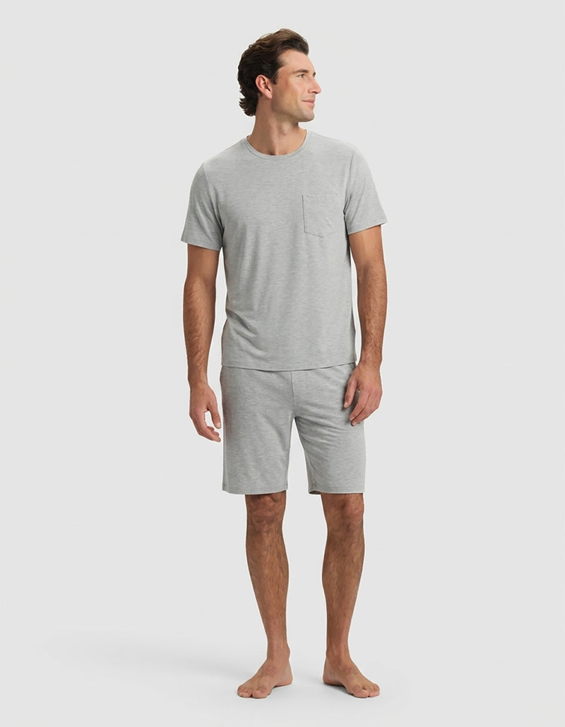 A man stands barefoot against a plain light background, wearing the Cozy Earth Men's Bamboo Stretch-Knit Short Sleeve Pajama Set in Heather Grey with a chest pocket and matching shorts, looking slightly to his left in a relaxed pose.
