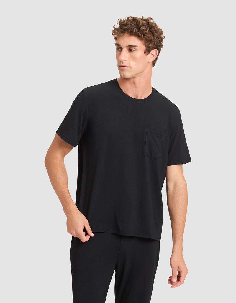 A young man with curly brown hair wears the Cozy Earth Men's Bamboo Stretch-Knit Lounge Tee in black, paired with matching pants. He stands against a light gray background, looking to his left with a neutral expression.