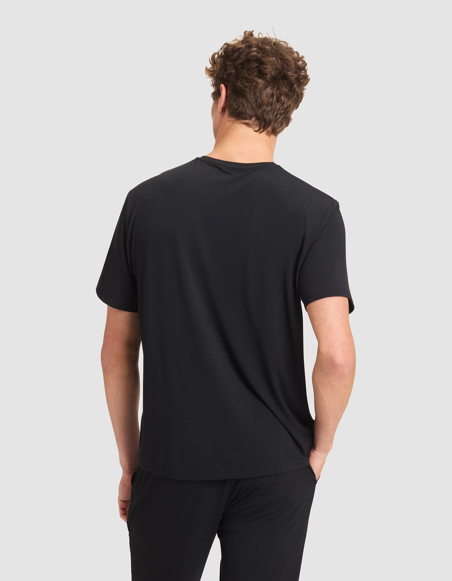 A person with short, curly brown hair stands with their back to the camera, wearing a Cozy Earth Men's Bamboo Stretch-Knit Lounge Tee and black pants against a light gray background. 