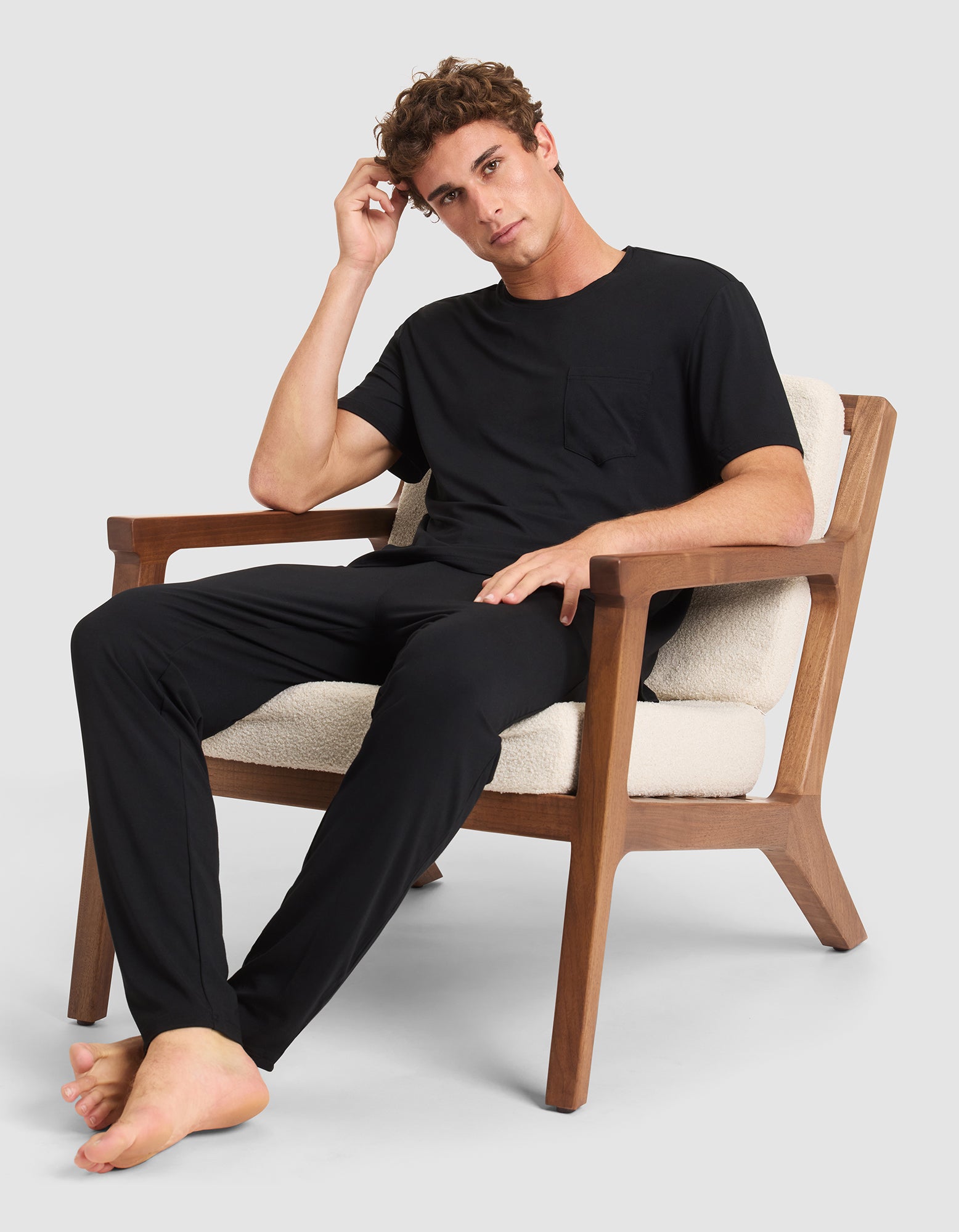 A young man in a Cozy Earth Men's Bamboo Stretch-Knit Lounge Tee and black pants sits barefoot on a wooden armchair with cream cushions, resting one hand on his head and wearing a neutral expression. 