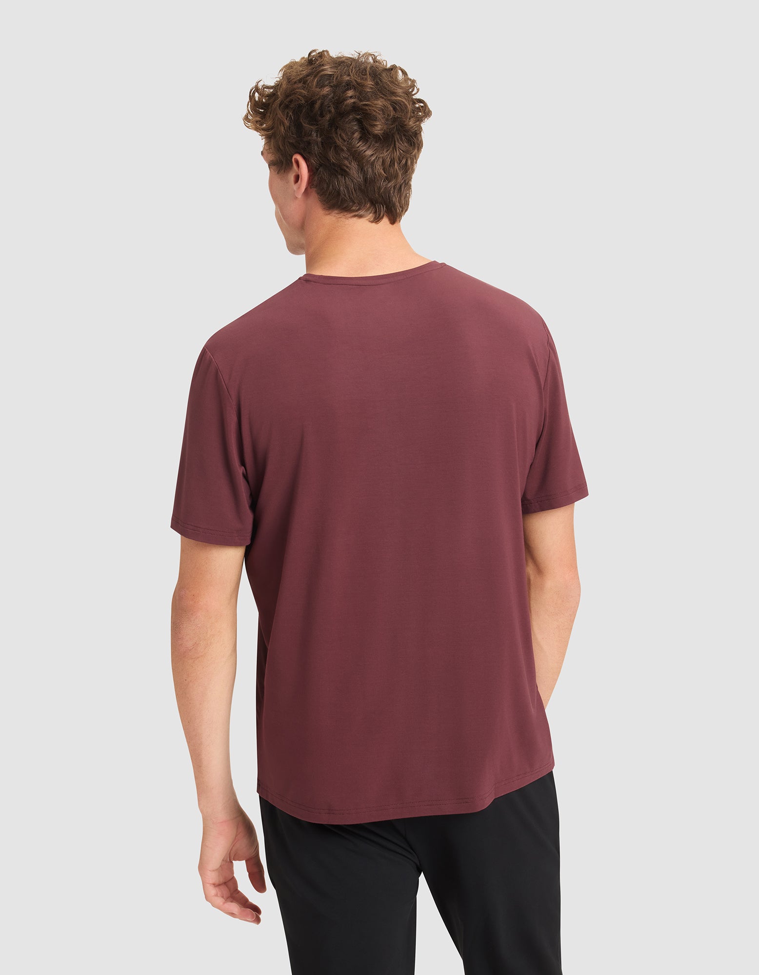 A man with curly brown hair is seen from behind, wearing the Cozy Earth Men's Bamboo Stretch-Knit Lounge Tee in maroon and black pants, standing against a plain light gray background.