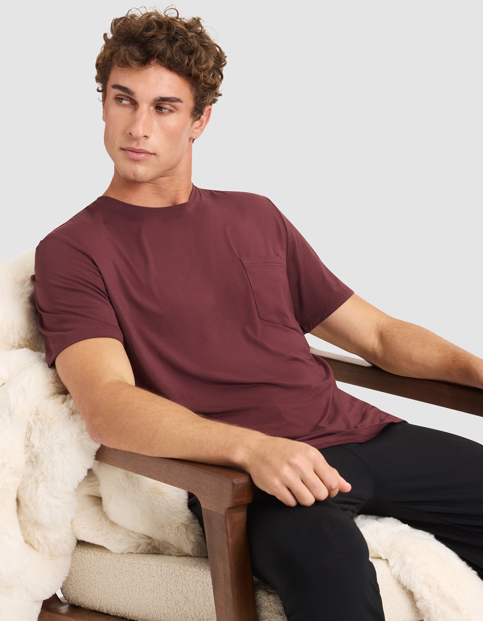 A young man with curly brown hair sits on a wooden armchair draped with a white fur throw, wearing Cozy Earth’s Men's Bamboo Stretch-Knit Lounge Tee in maroon and black pants, looking to his left with a neutral expression.