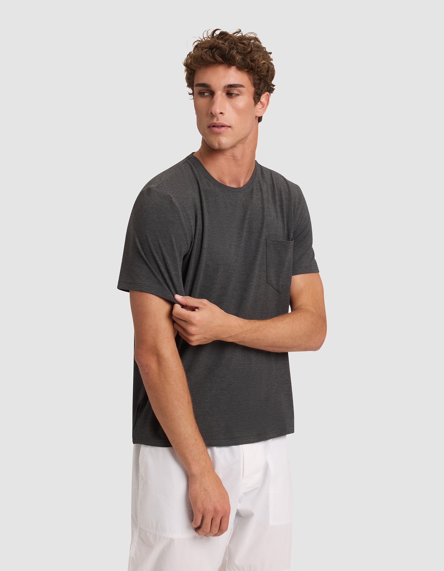 A young man with curly hair wears a Cozy Earth Men's Bamboo Stretch-Knit Lounge Tee in dark gray and white shorts, standing against a light gray background while holding the hem of his shirt and looking to the side. |Color:Charcoal
