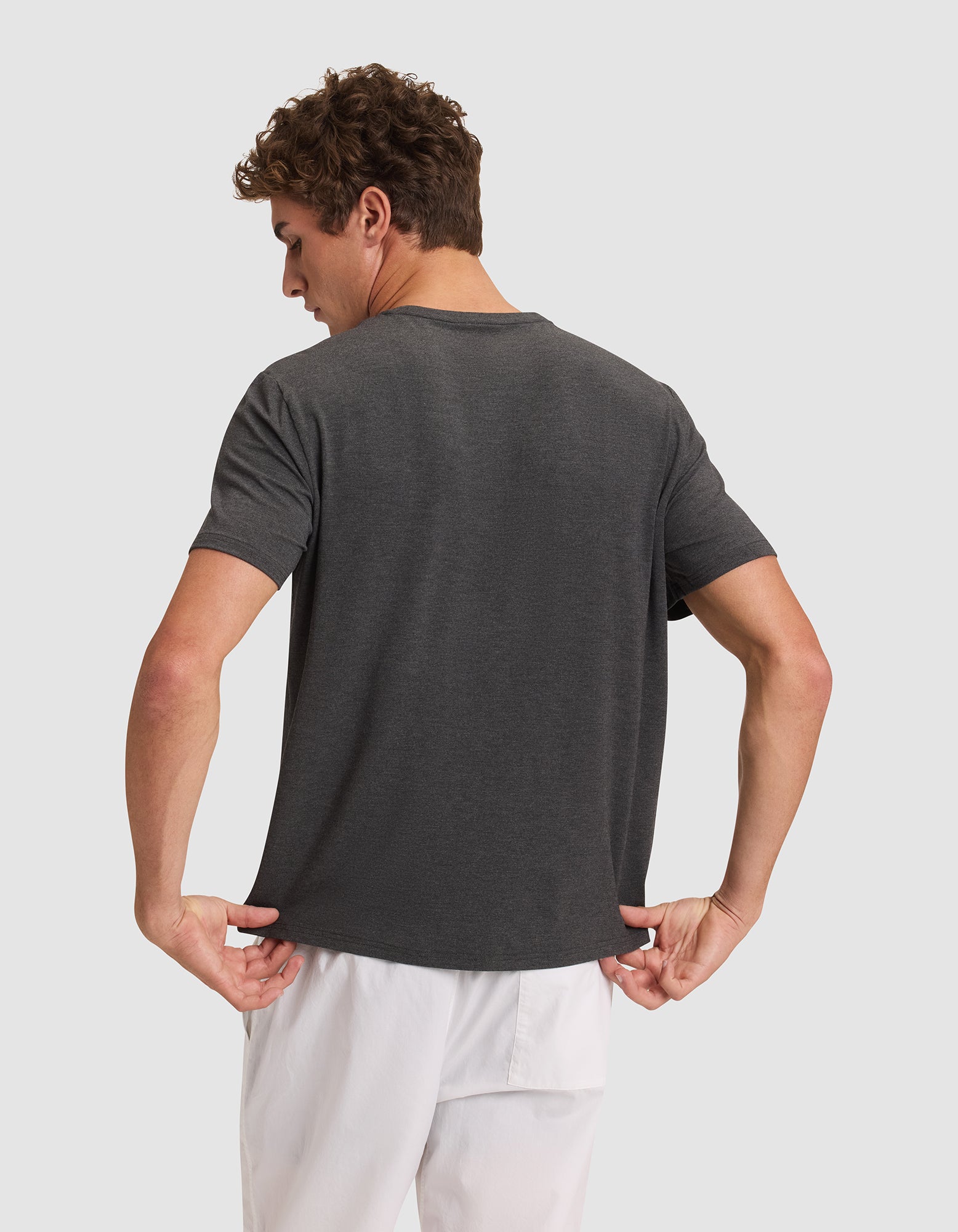 A man faces away from the camera, wearing the Cozy Earth Men's Bamboo Stretch-Knit Lounge Tee in dark gray and white shorts. He holds the bottom of his tee with both hands against a plain, light background.