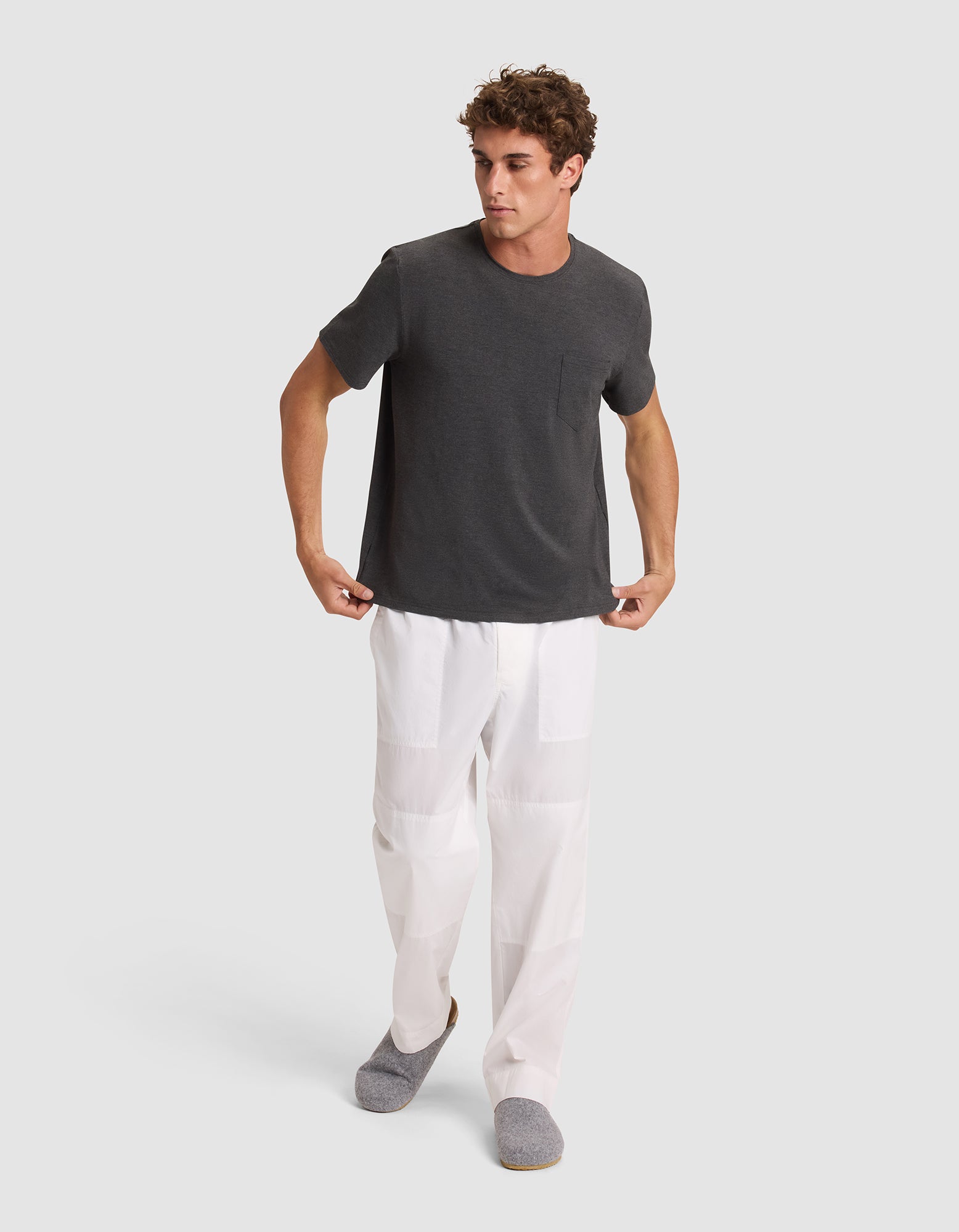 A man wearing the Cozy Earth Men's Bamboo Stretch-Knit Lounge Tee in dark gray stands against a light gray background, looking to his left with his arms slightly out to his sides.