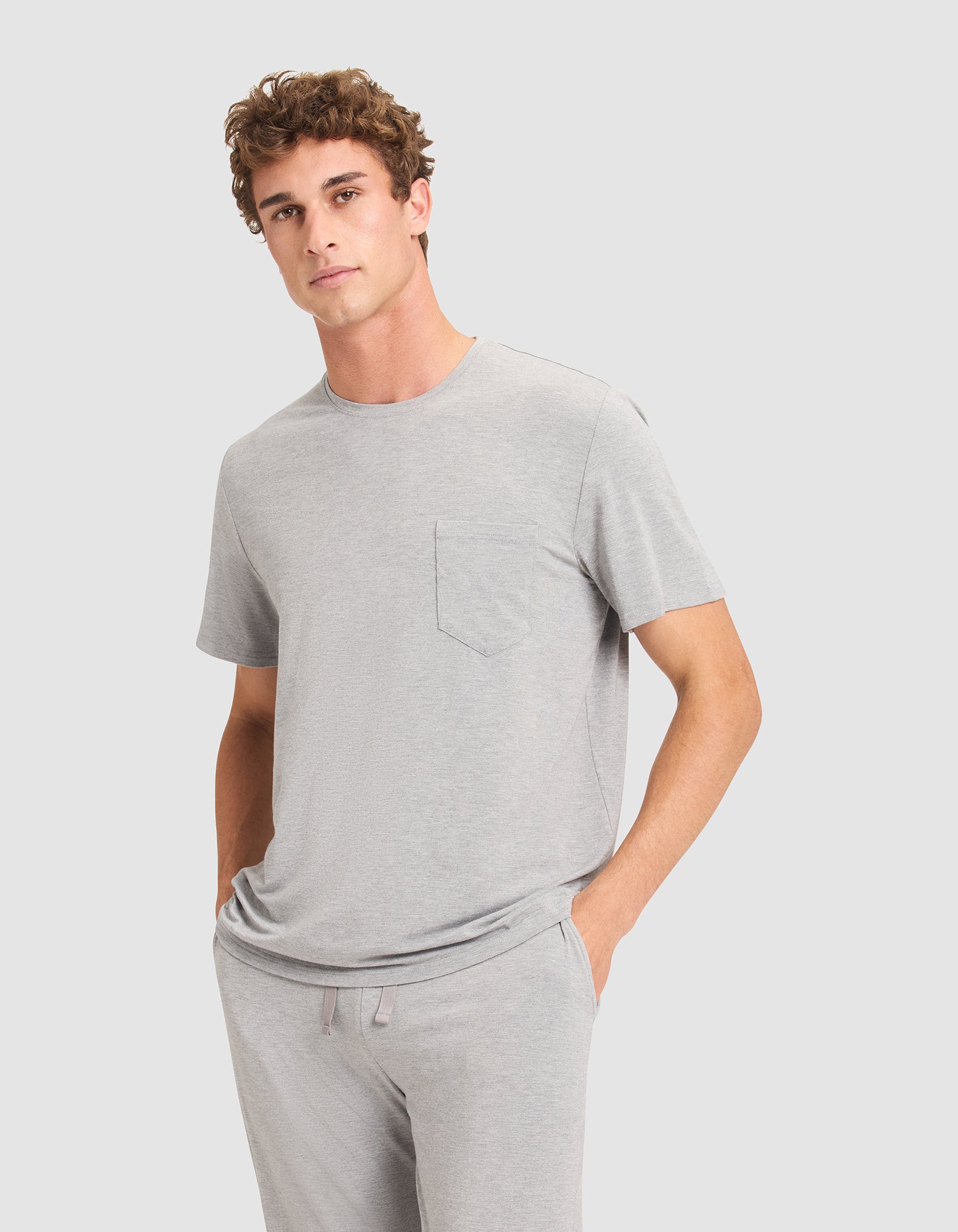 A young man with short curly hair stands with his hands in his pockets, wearing Cozy Earth's Men's Bamboo Stretch-Knit Lounge Tee in light gray and matching gray pants against a plain light background. 