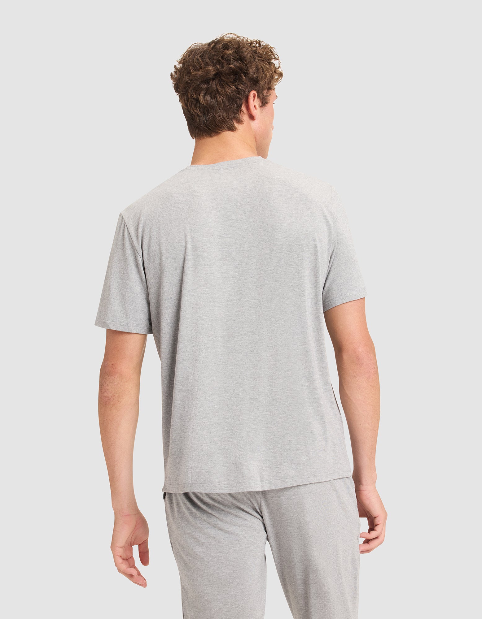 A man with curly hair, seen from behind, wears a light gray Cozy Earth Men's Bamboo Stretch-Knit Lounge Tee and matching pants against a plain, light background. |Color:Heather Grey
