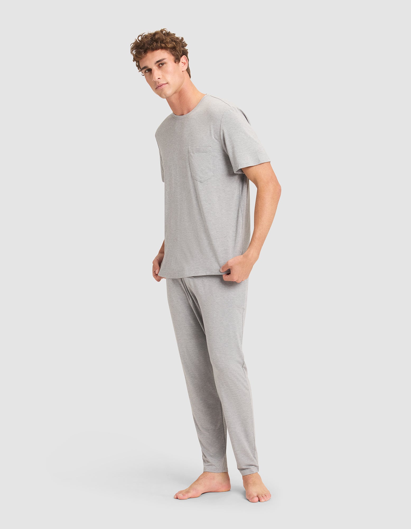 A man stands barefoot against a plain background, wearing the Cozy Earth Men's Bamboo Stretch-Knit Lounge Tee in light gray with a chest pocket, paired with matching long pajama pants. 