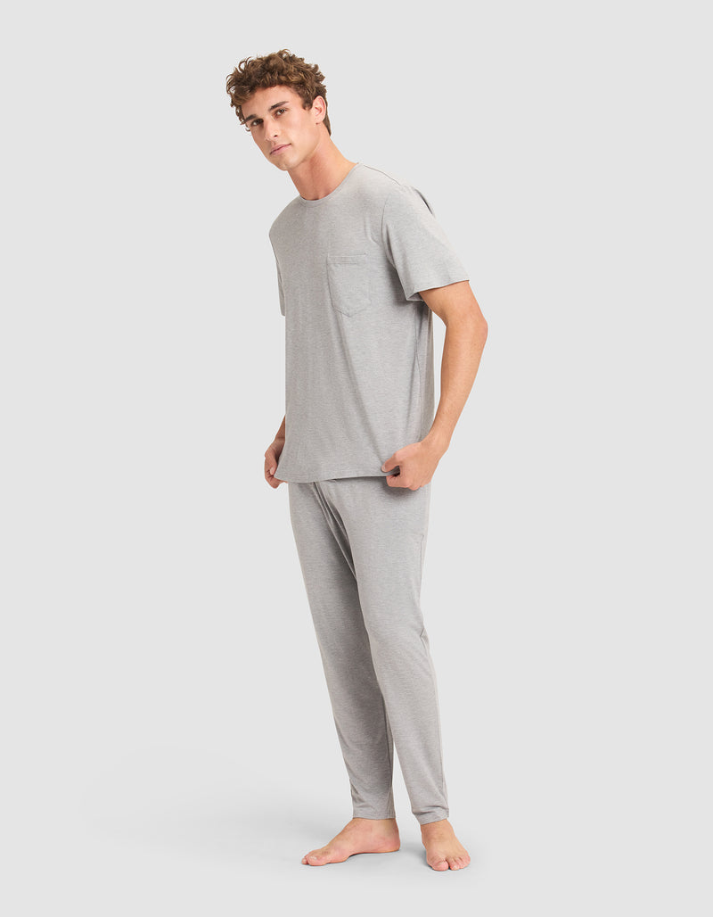 A man stands barefoot against a plain background, wearing the Cozy Earth Men's Bamboo Stretch-Knit Lounge Tee in light gray with a chest pocket, paired with matching long pajama pants.