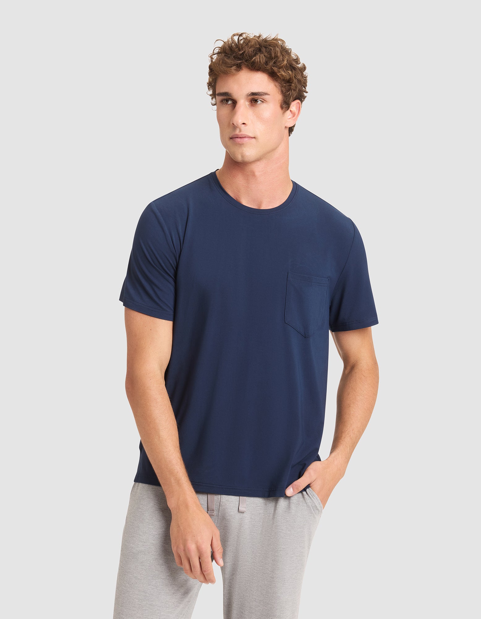 A young man with curly hair models the Cozy Earth Men's Bamboo Stretch-Knit Lounge Tee in navy blue with short sleeves and pocket, paired with light gray pants, standing against a plain light gray background.  