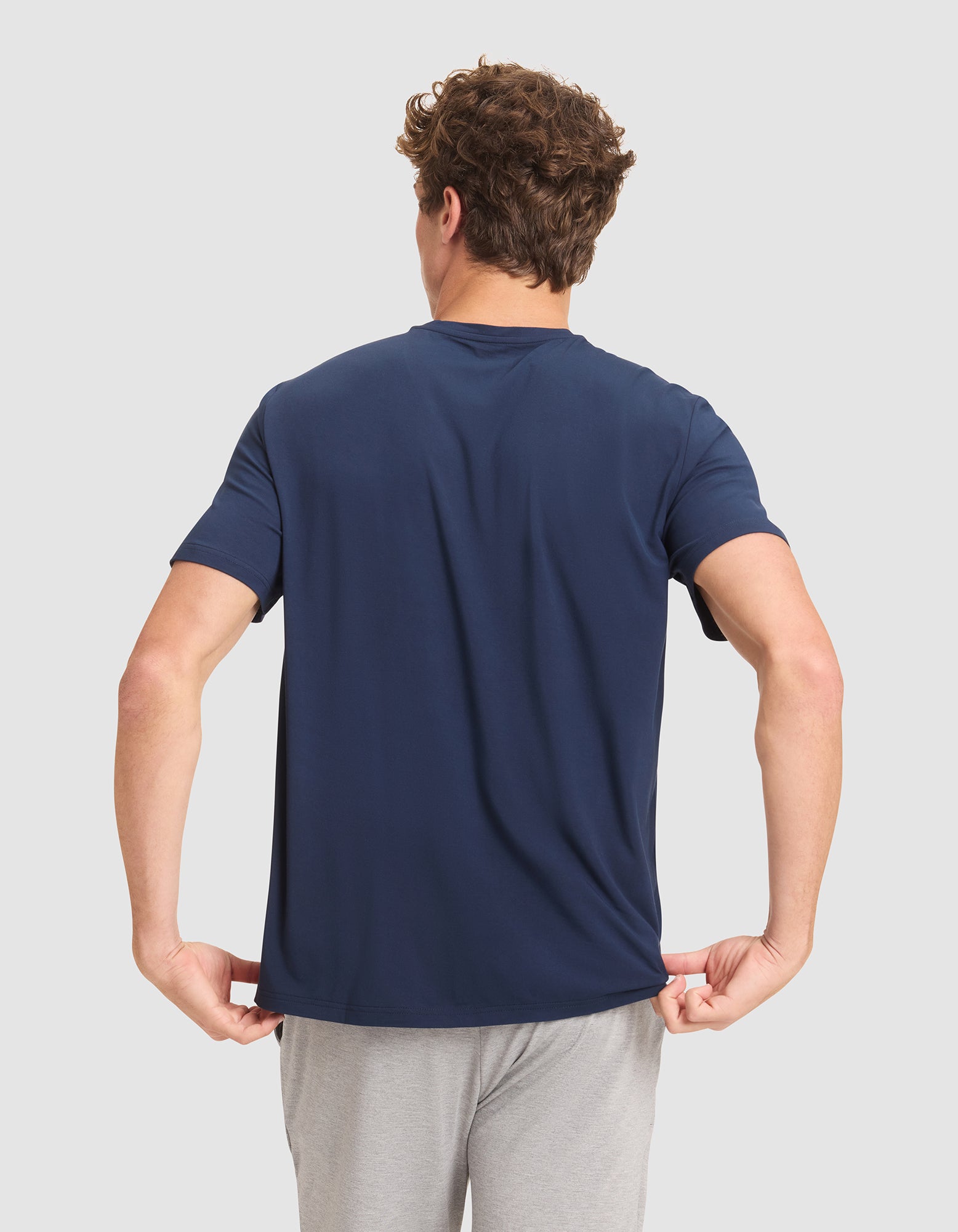 A man with curly hair faces away from the camera, wearing Cozy Earth's Men's Bamboo Stretch-Knit Lounge Tee in navy blue and light gray shorts, holding the hem of his shirt with both hands against a plain background.  