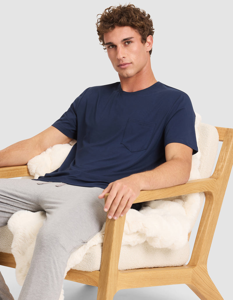A man with curly brown hair sits relaxed in a wooden chair with white cushions, wearing the Cozy Earth Men's Bamboo Stretch-Knit Lounge Tee in navy blue and light gray pants, one arm resting on the chair.