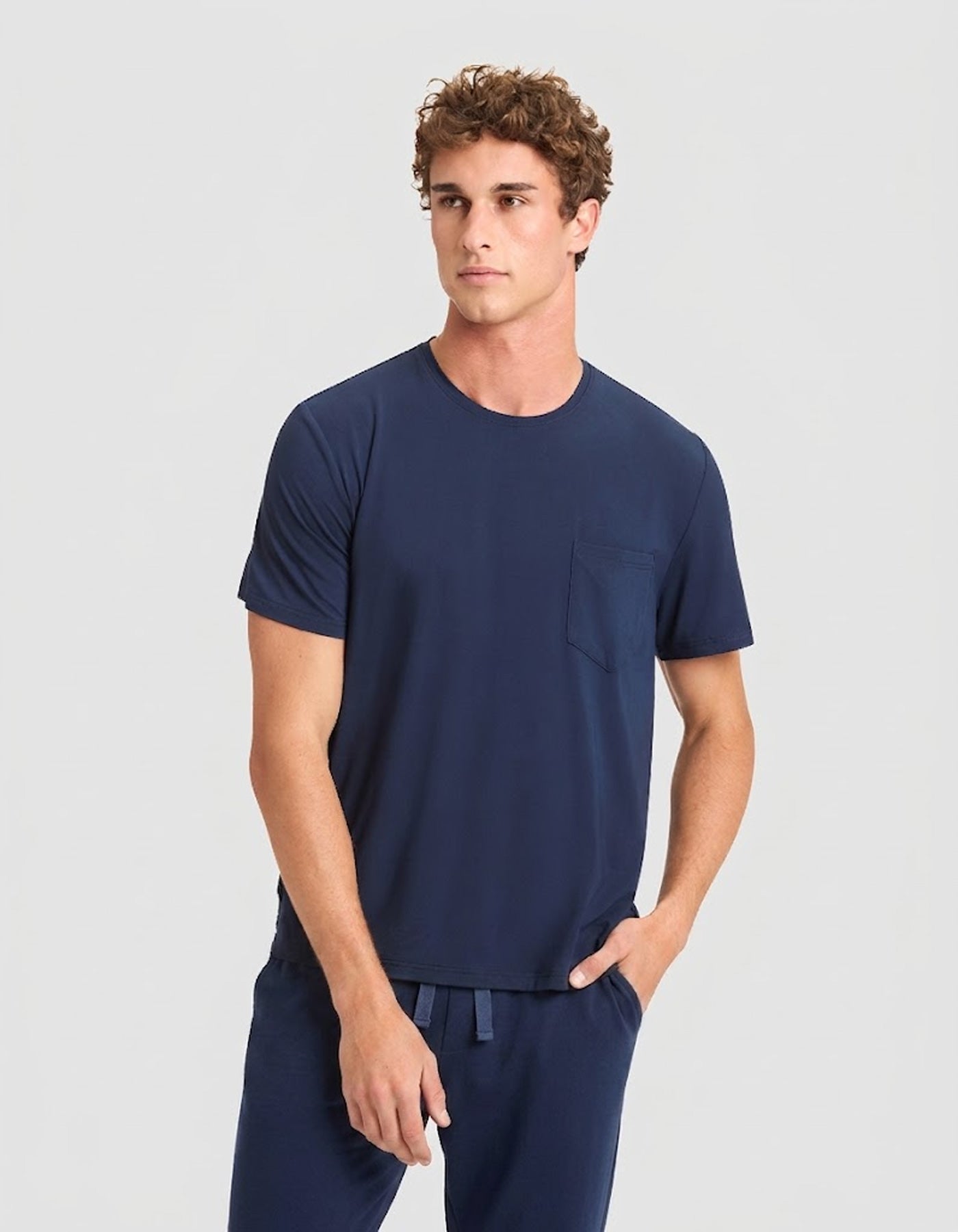 A young man with short curly hair models the Cozy Earth Men's Bamboo Stretch-Knit Lounge Tee & Pant Pajama Set in navy, featuring a short-sleeve chest-pocket tee and matching drawstring pants, against a plain light gray background.