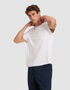 A young man stands against a plain light gray background, wearing Cozy Earth’s Men’s Bamboo Stretch-Knit Lounge Tee in white with a chest pocket and dark pants, his hands resting on the back of his neck.  |Color:White