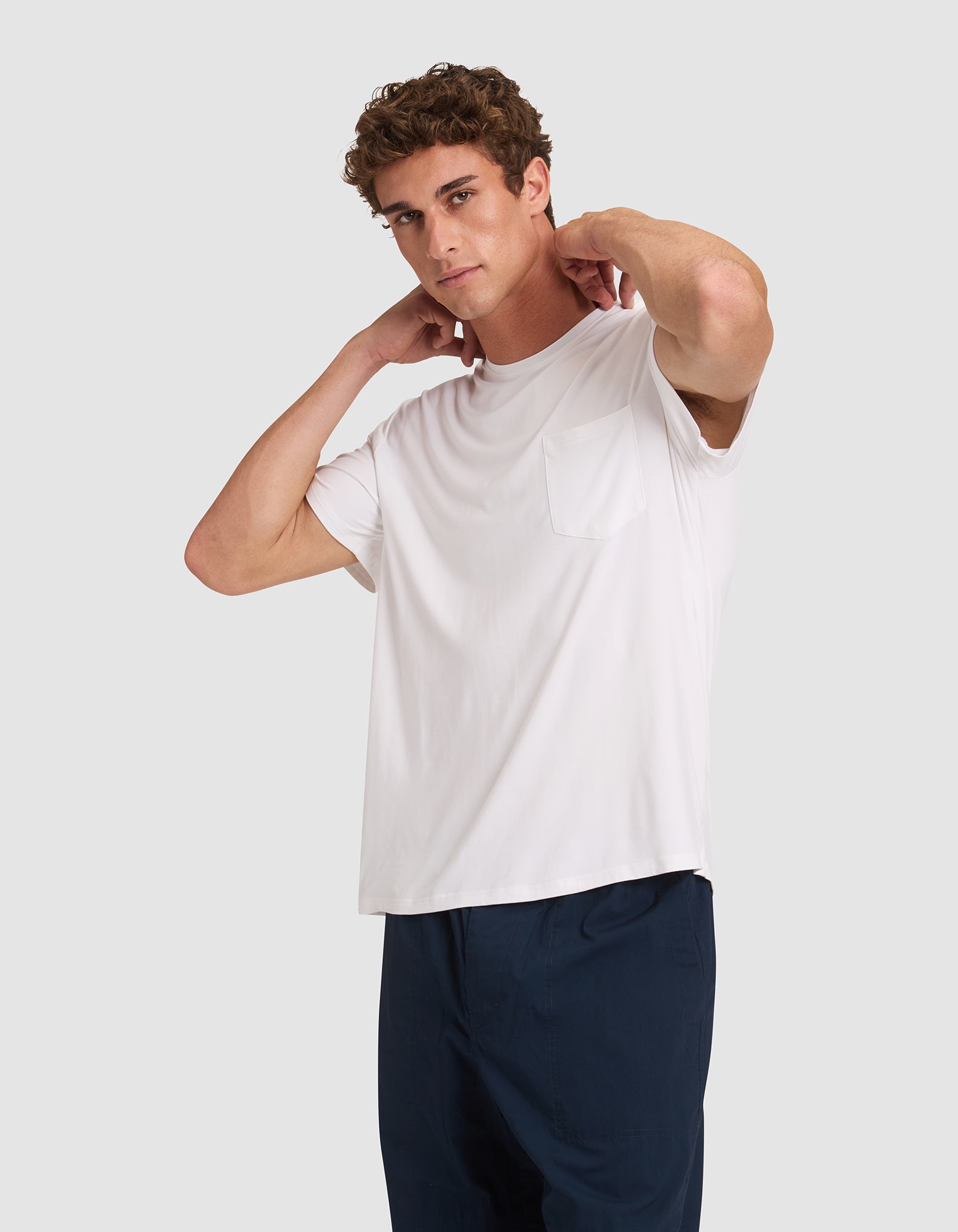 A young man stands against a plain light gray background, wearing Cozy Earth’s Men’s Bamboo Stretch-Knit Lounge Tee in white with a chest pocket and dark pants, his hands resting on the back of his neck.  