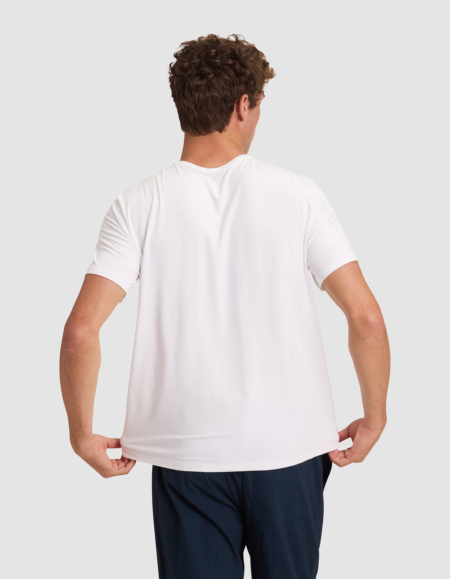 A man with curly brown hair, seen from behind, wears a plain white Men's Bamboo Stretch-Knit Lounge Tee by Cozy Earth and dark pants. He pulls the sides of his shirt slightly outward against a light gray background. 