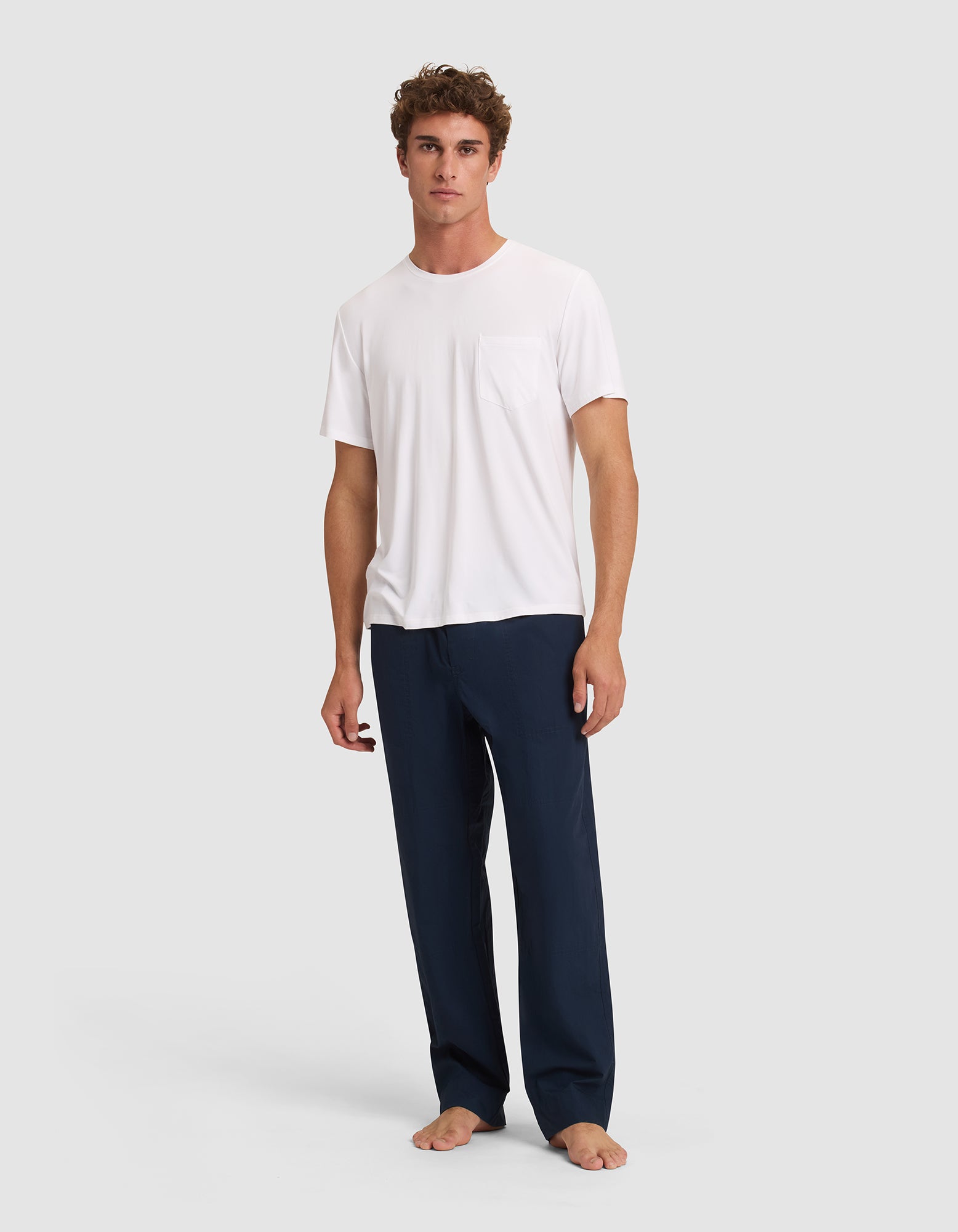 A person stands barefoot against a plain background, wearing the Cozy Earth Men's Bamboo Stretch-Knit Lounge Tee in white and loose-fitting navy blue pants.  