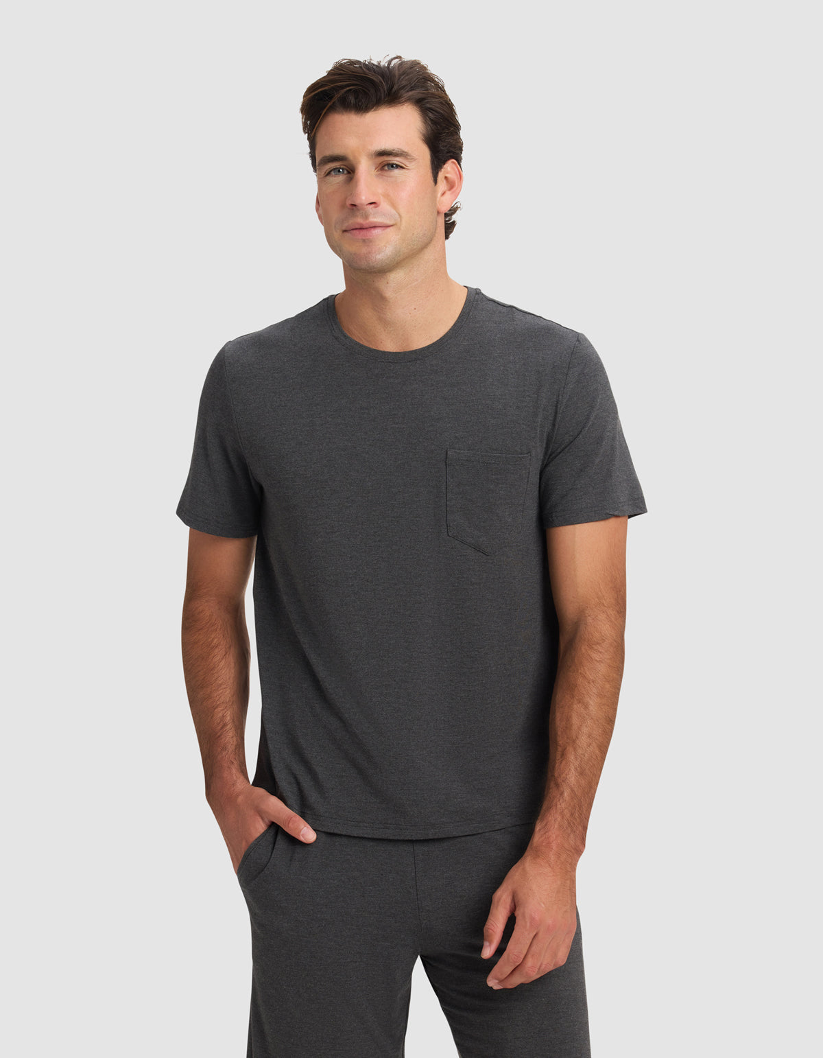 A man wearing the Cozy Earth Men's Bamboo Stretch-Knit Lounge Tee in charcoal gray, featuring short sleeves and a chest pocket, stands against a light gray background with one hand in his pocket, smiling slightly.|Color:Charcoal