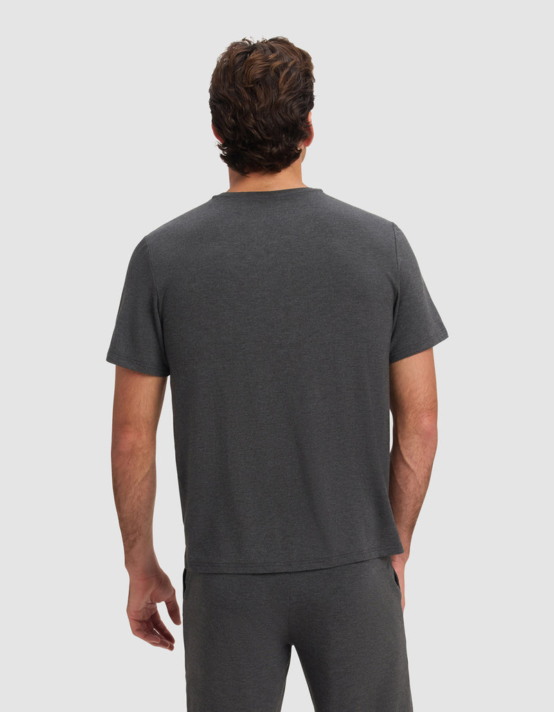 A man with short, wavy brown hair is shown from the back wearing Cozy Earth's Men's Bamboo Stretch-Knit Lounge Tee in dark gray, paired with matching pants, against a plain light gray background.