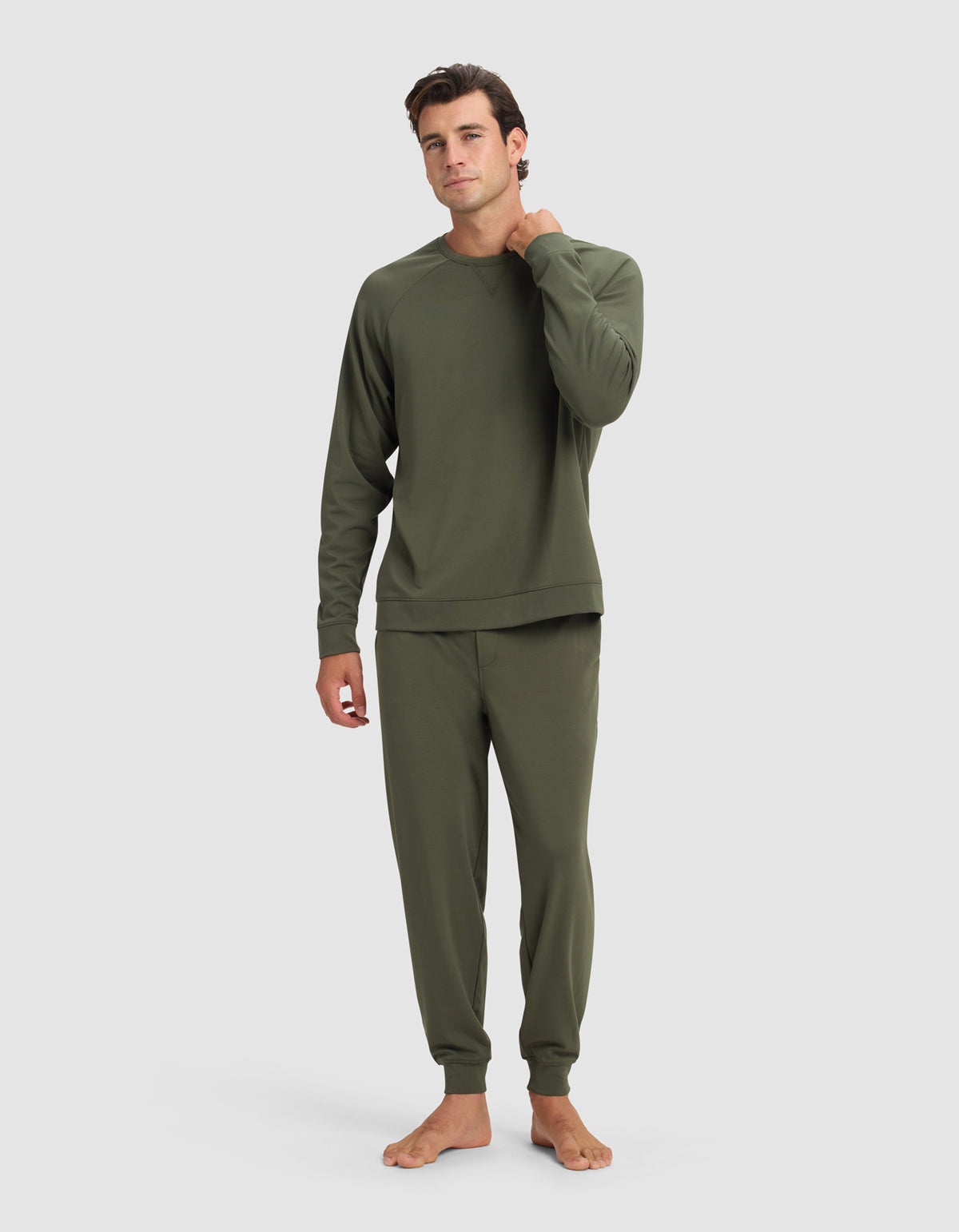 A man stands barefoot against a plain light background, wearing the Cozy Earth Men's Brushed Bamboo Jogger Set in olive green. He poses with one hand at his neck, looking relaxed.