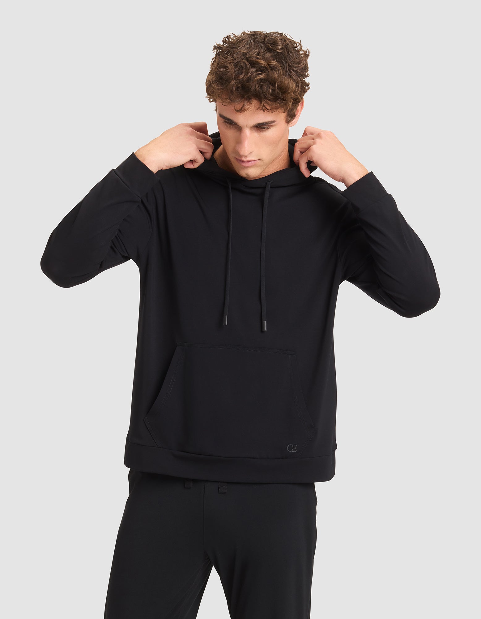 A young man with curly hair adjusts the hood of a black Cozy Earth Men's Brushed Bamboo Hoodie, paired with black pants, while standing against a plain light gray background. 