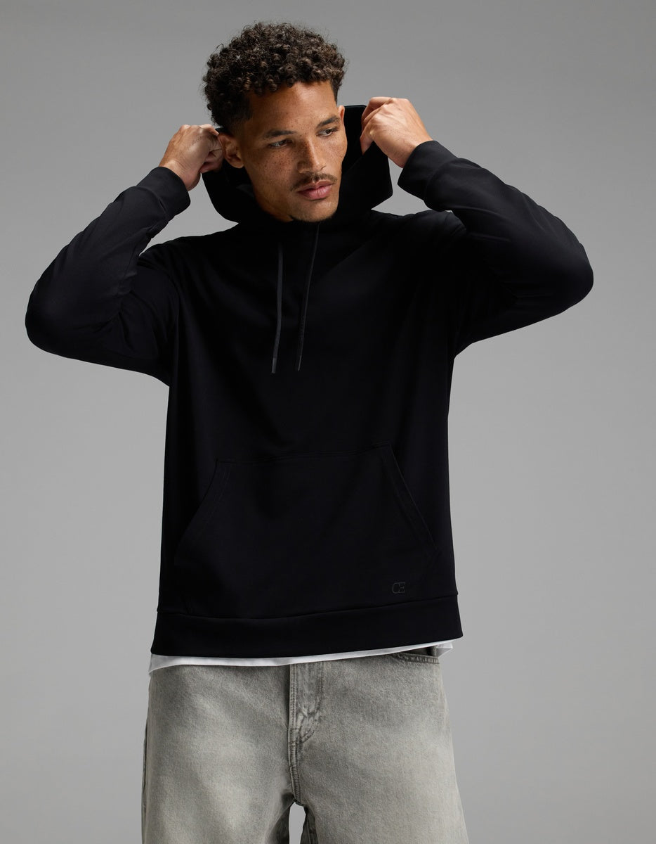 A man wearing the Cozy Earth Men’s Brushed Bamboo Hoodie in black and light gray jeans adjusts the hood while looking slightly down against a plain gray background.