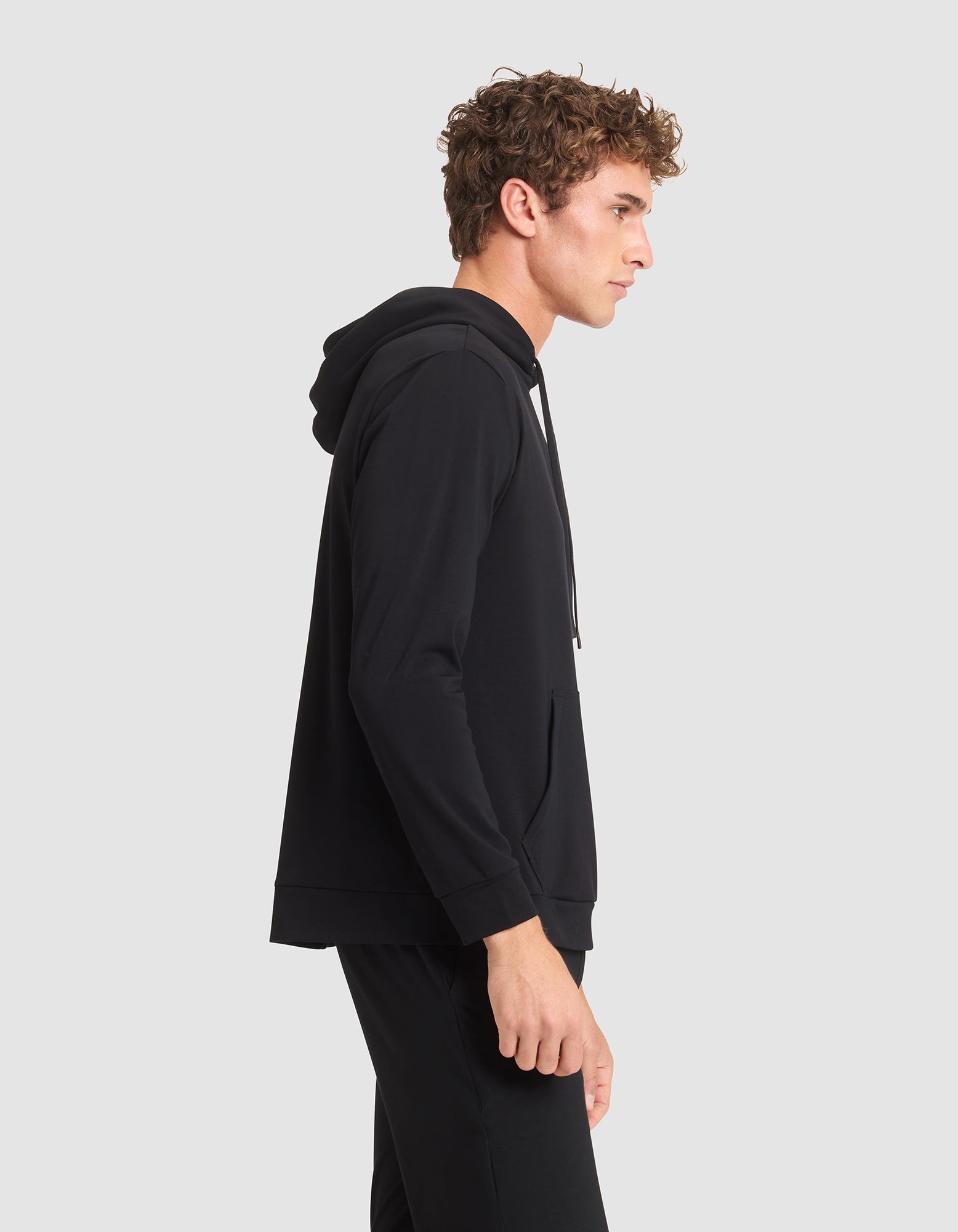A young man with curly hair stands in profile, wearing Cozy Earth's Men's Brushed Bamboo Hoodie and black pants against a plain light gray background. 