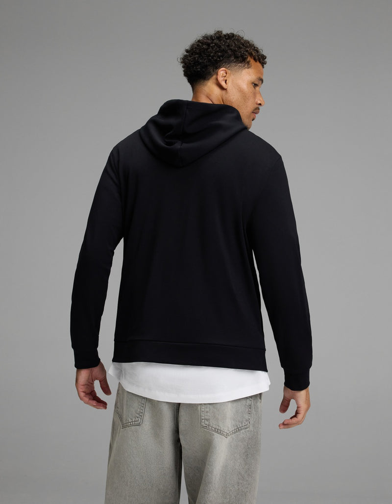A man stands facing away, wearing the Cozy Earth Men’s Brushed Bamboo Hoodie in black over a white shirt and light gray jeans, set against a plain gray background.