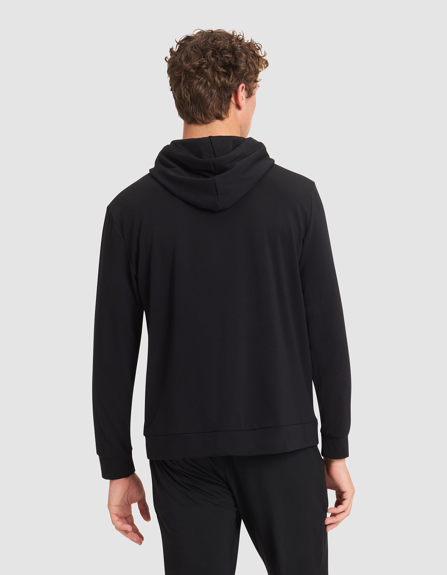 A man with curly hair is seen from behind wearing a Cozy Earth Men's Brushed Bamboo Hoodie in black, paired with black pants, set against a plain light gray background. 